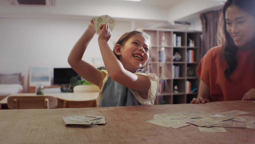 A joyful little girl excitedly celebrates winning a card game with a woman in their cozy living room, captured in slow motion