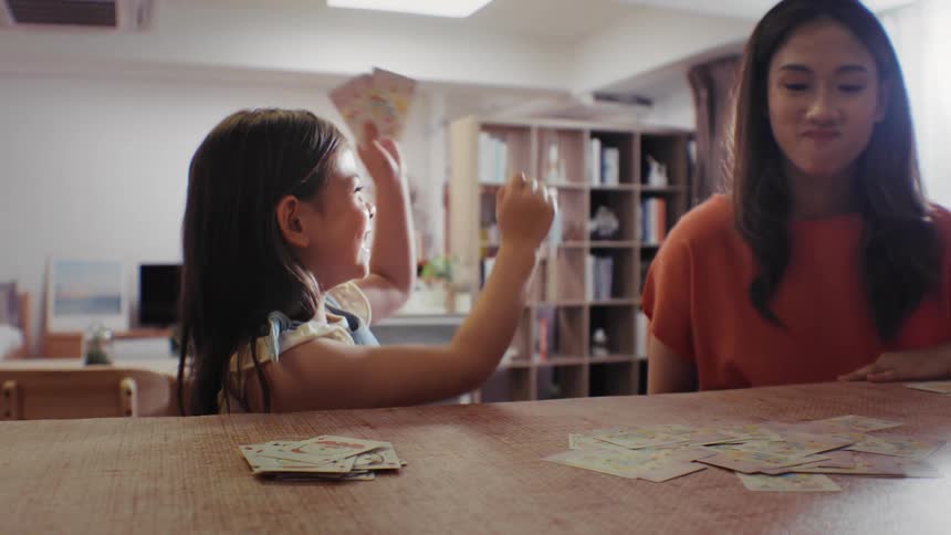 A joyful little girl excitedly celebrates winning a card game with a woman in their cozy living room, captured in slow motion