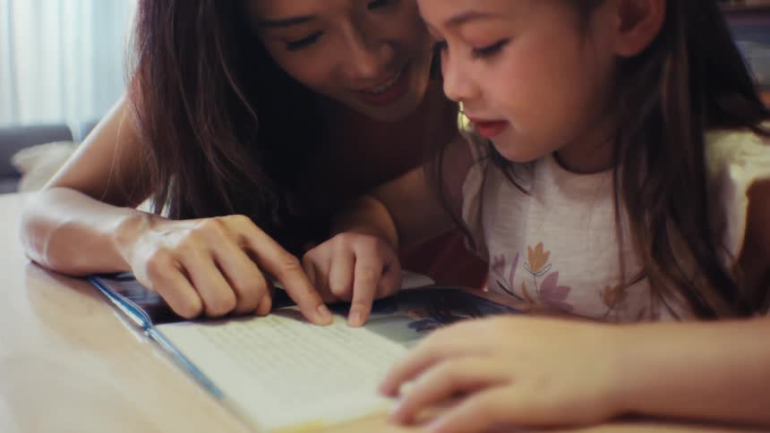 A loving mother helps her young daughter learn to read as they trace the words in a book together at home