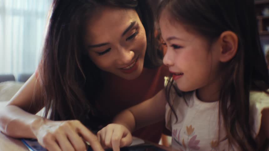 A loving mother helps her young daughter learn to read as they trace the words in a book together at home