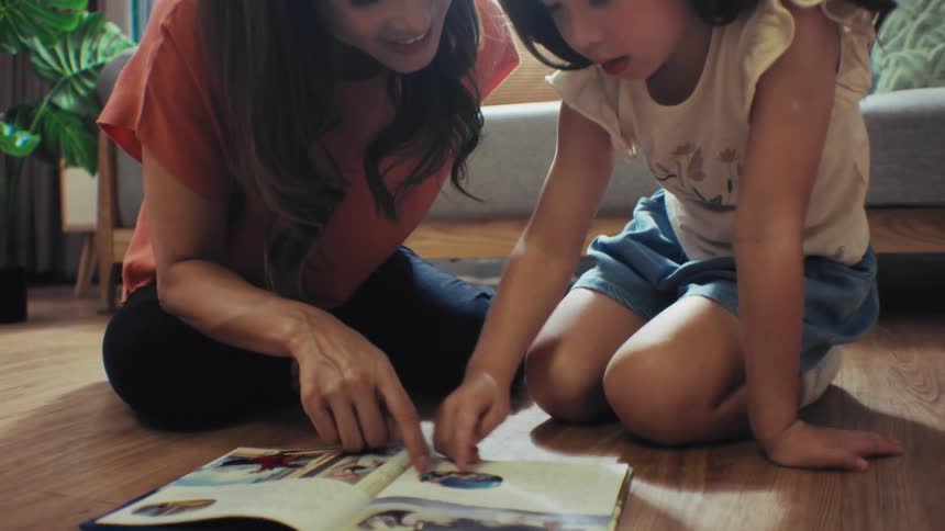 An adorable young girl laughs joyfully while reading a book with her mother on the floor of their bright living room