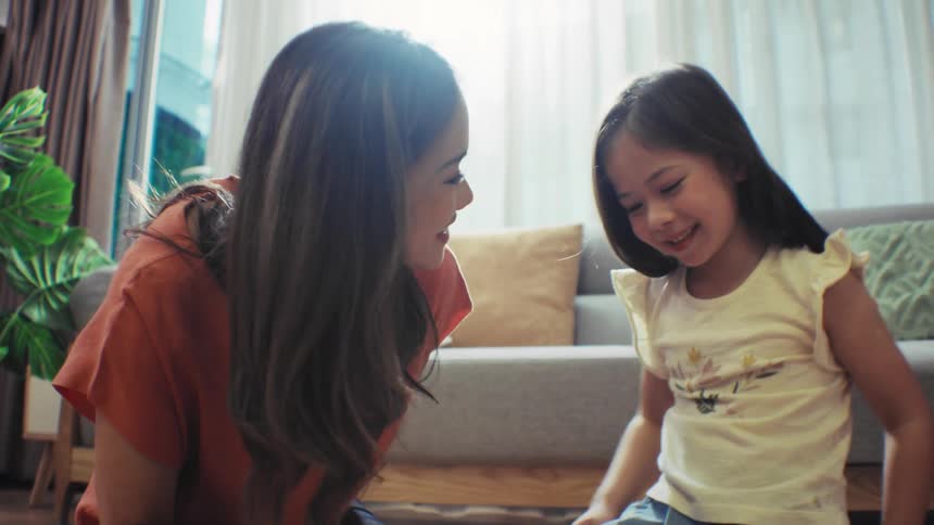 An adorable young girl laughs joyfully while reading a book with her mother on the floor of their bright living room