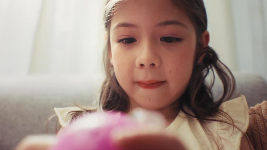 A close-up of a cute young girl with long hair intently focusing on a pink toy in her hands in a brightly lit room