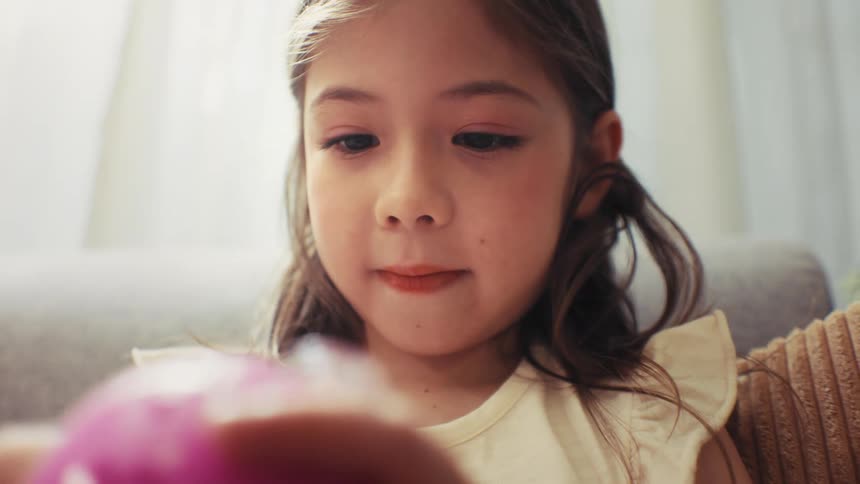 A close-up of a cute young girl with long hair intently focusing on a pink toy in her hands in a brightly lit room