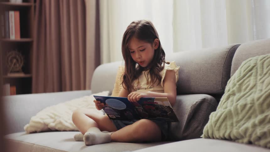 A focused young girl sits comfortably on a sofa in a sunlit living room, completely absorbed in reading a colorful picture book