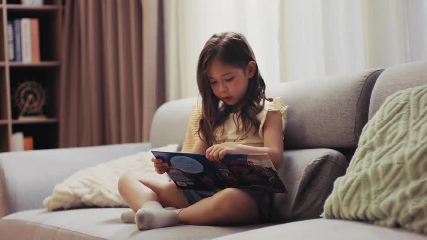 A focused young girl sits comfortably on a sofa in a sunlit living room, completely absorbed in reading a colorful picture book