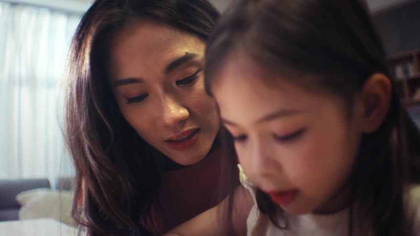 An intimate close-up shows a mother gently encouraging her focused daughter during an activity at home