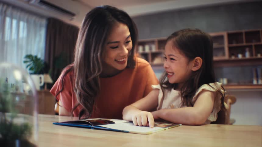A loving mother and her happy young daughter read a book together at the dining table, smiling and sharing a warm family moment