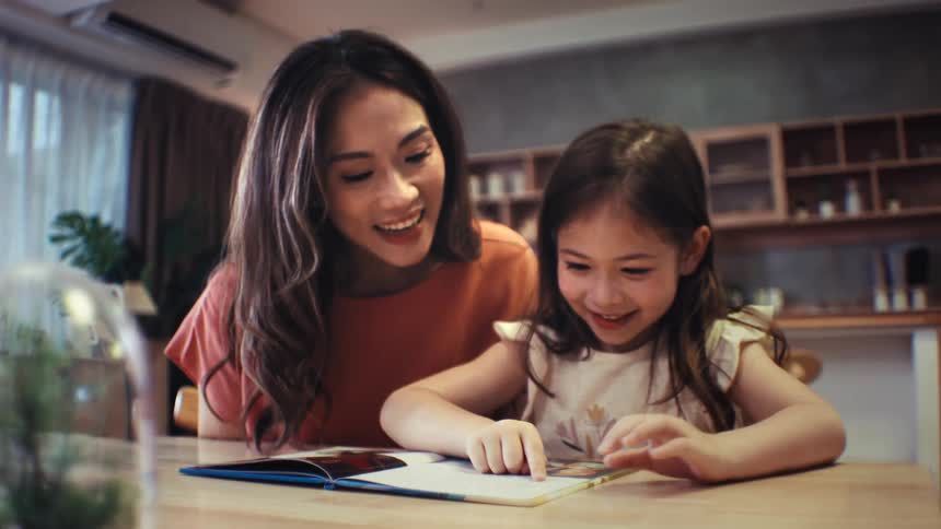 A loving mother and her happy young daughter read a book together at the dining table, smiling and sharing a warm family moment