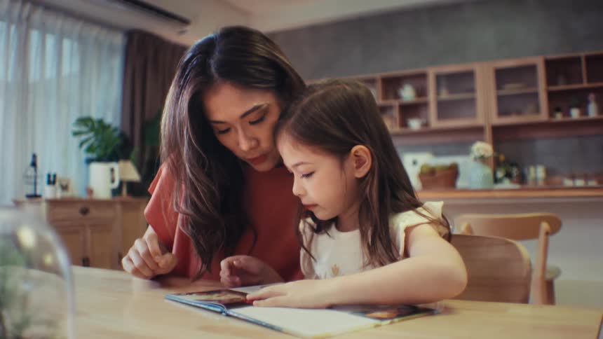 A loving mother helps her young daughter learn to read a book while sitting together at the table in their cozy home