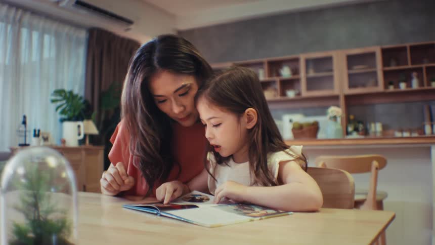 A loving mother helps her young daughter learn to read a book while sitting together at the table in their cozy home
