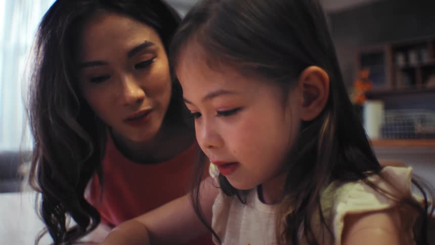 A loving Asian mother proudly listens as her young daughter practices reading a book at the table