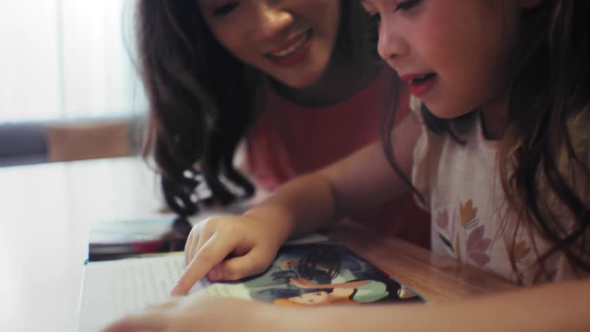 A loving Asian mother proudly listens as her young daughter practices reading a book at the table