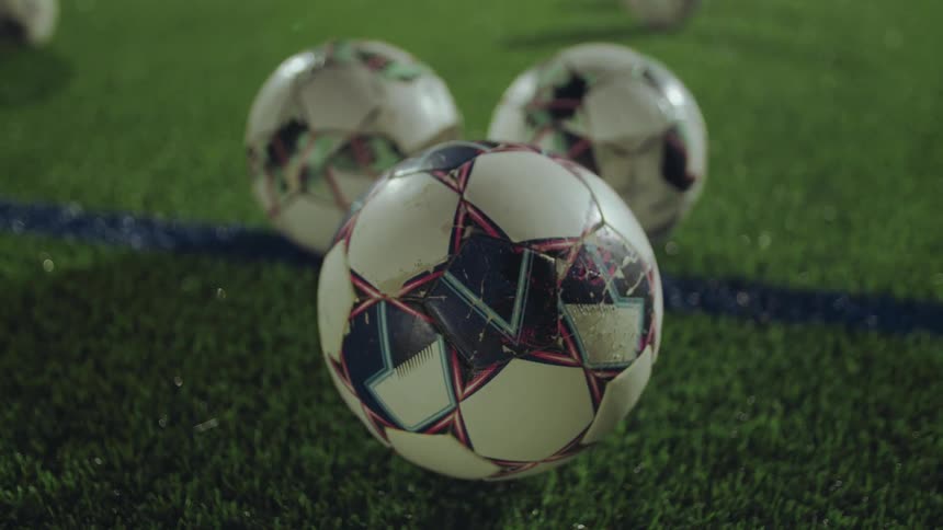 Close-up of three weathered soccer balls resting on wet artificial turf at night as a light rain falls