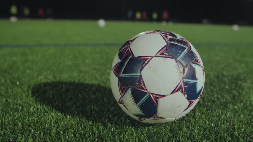 A close-up of a soccer ball on a green turf field at night with players practicing in the background