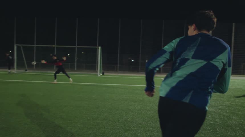 From behind a player in slow motion as they run up and kick a soccer ball, scoring a goal at night