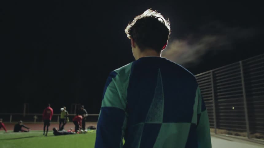 A young male athlete walks onto the field for a cold night practice, his breath visible under the stadium lights