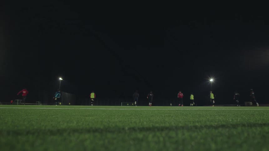 Low angle, out-of-focus view of a team of athletes running across a green turf field during a nighttime training session