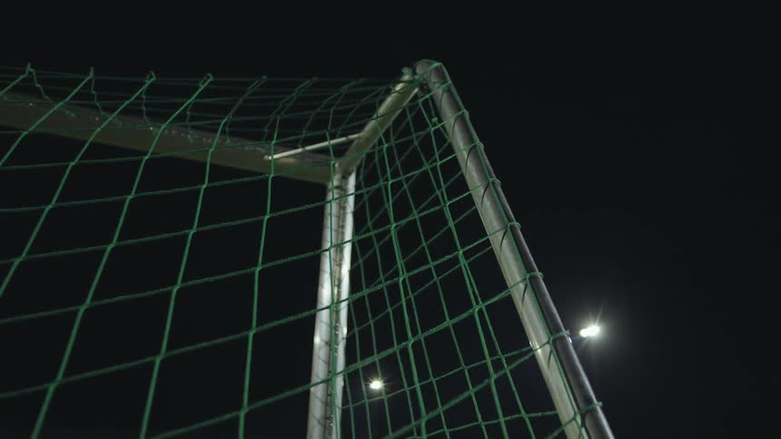 A soccer ball hits the back of the net as a goal is scored on a field at night under stadium lights