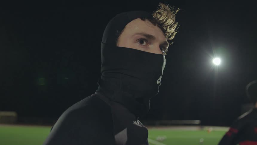 A young male athlete wearing a black balaclava stands with an intense, focused expression on a sports field at night