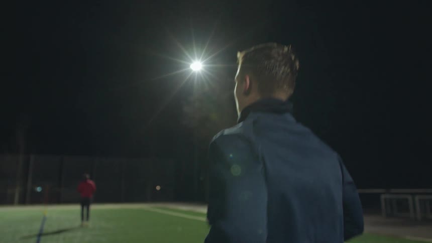 A focused athlete runs in slow motion on a turf field at night under a bright stadium light