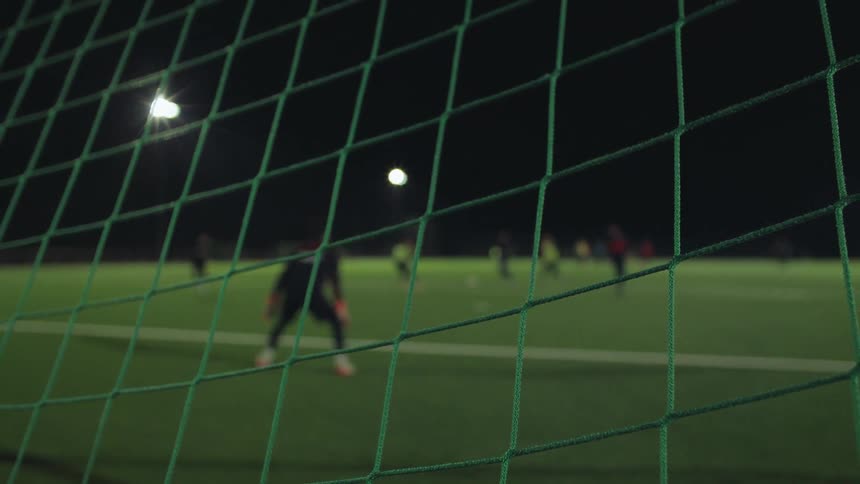Close up on a soccer goal net with a team training on the illuminated field in the background at night