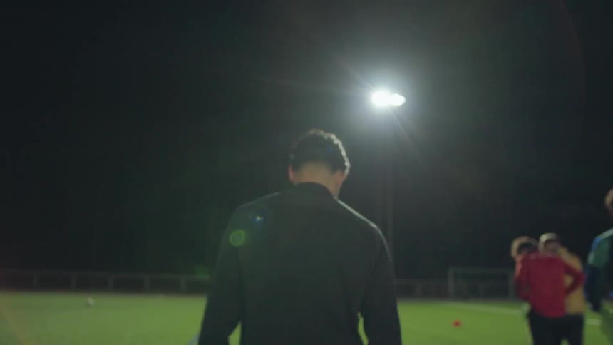 A male coach jogs across a soccer field during a team training session at night under bright stadium lights