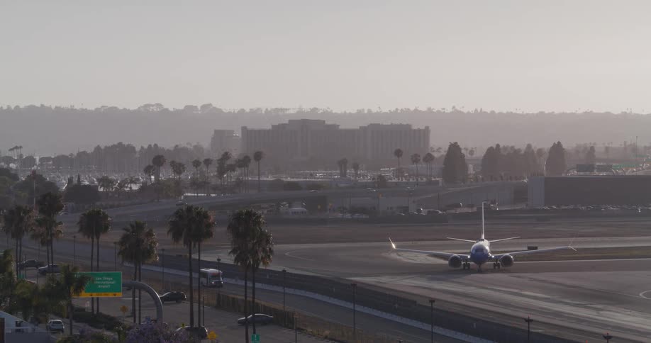 A passenger airplane sits on the tarmac at San Diego International Airport with highway traffic and palm trees in the foreground on a hazy morning