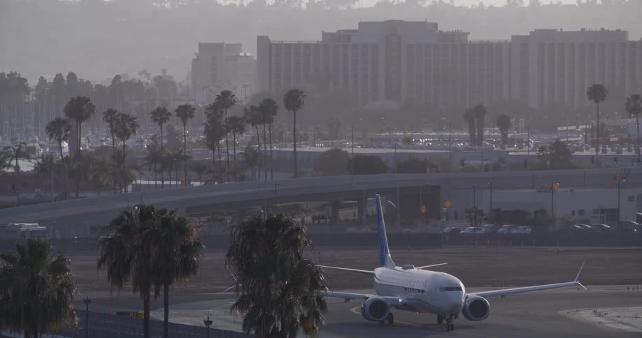 A United Airlines plane taxis on a hazy airport runway surrounded by palm trees and city buildings