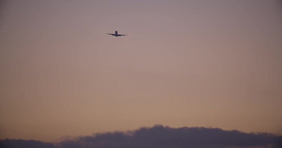 A passenger airplane with blinking lights flies away into the distance against a warm twilight sky