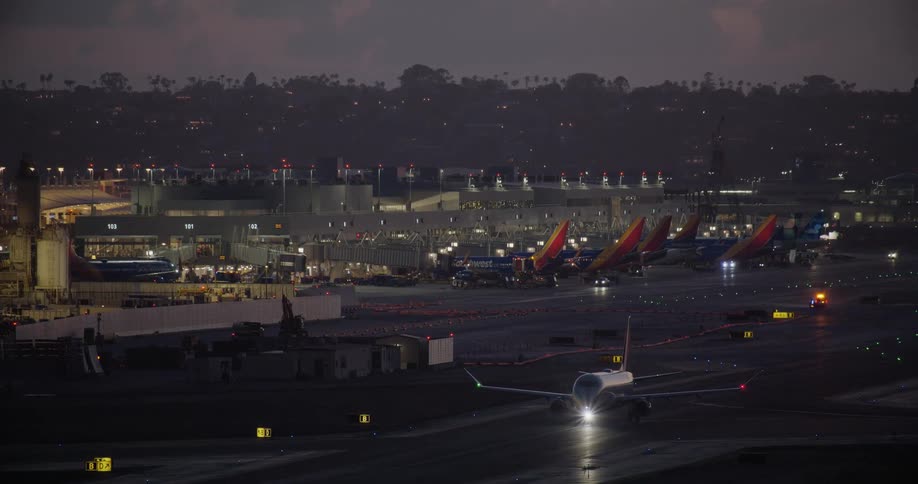 An airplane with its bright headlights on taxis down the runway in front of a busy airport terminal at twilight