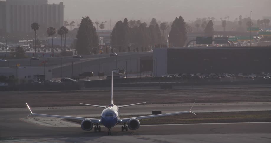 A head-on static shot of a commercial airplane on the tarmac at a hazy airport with traffic in the background