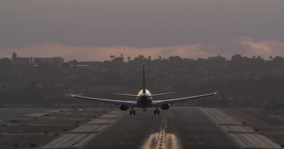 A commercial jetliner descends from a golden, cloudy sky to gracefully land on a city airport runway in slow motion