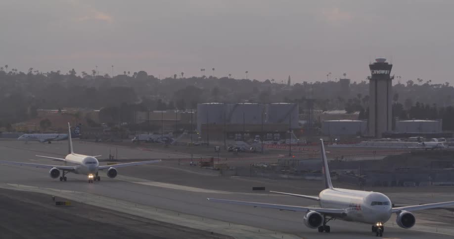 A cargo plane taxis on an airport runway at dawn, with another commercial jet and the air traffic control tower visible in the background