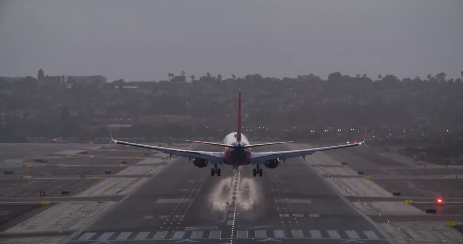 A commercial airliner seen from behind makes its final descent over a city before landing on an illuminated runway at dusk