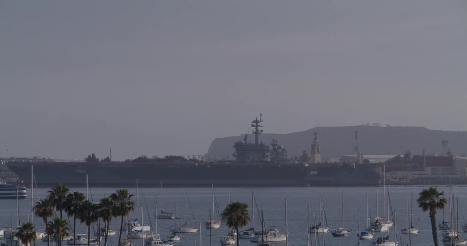 A static shot of an aircraft carrier docked in the San Diego harbor, with a marina of sailboats and palm trees in the foreground