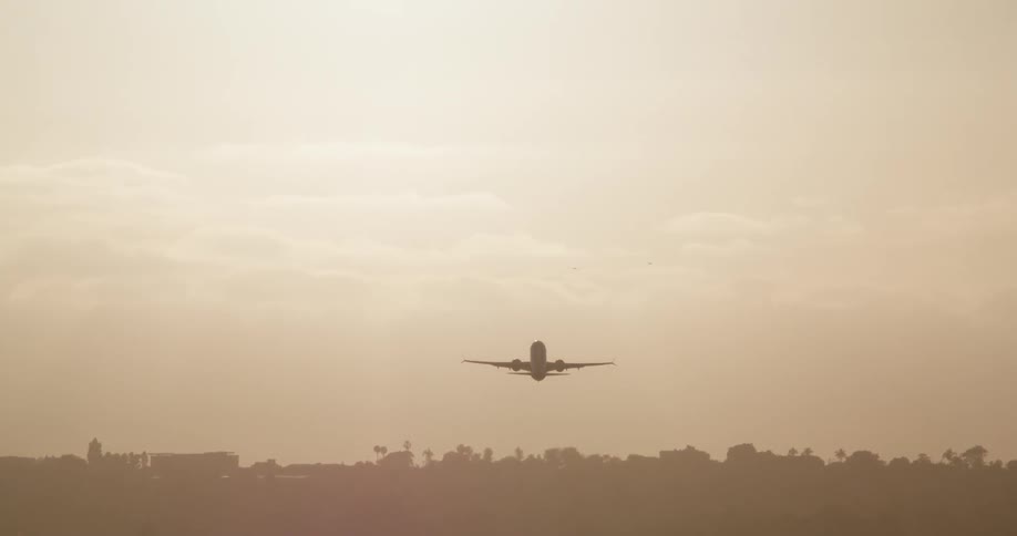 A passenger airplane takes off from a reflective runway during a hazy golden hour, ascending and flying into the bright sun