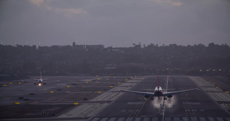 A commercial airliner makes a dramatic landing on an illuminated runway at dusk with the city in the background