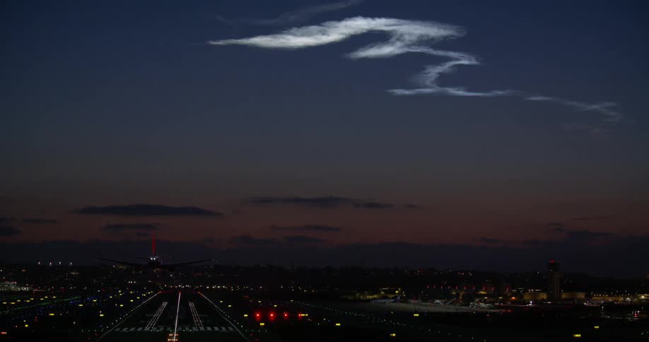 A passenger jet descends for landing on a brightly lit airport runway at night under a unique snake-like cloud formation
