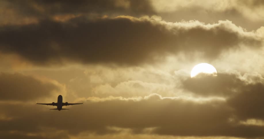 A silhouetted airplane takes off and flies away into a beautiful, cloudy, golden sky at sunrise