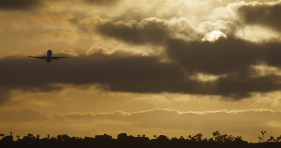 A passenger jet is silhouetted as it takes off and flies into a dramatic golden sky filled with dark clouds at sunrise