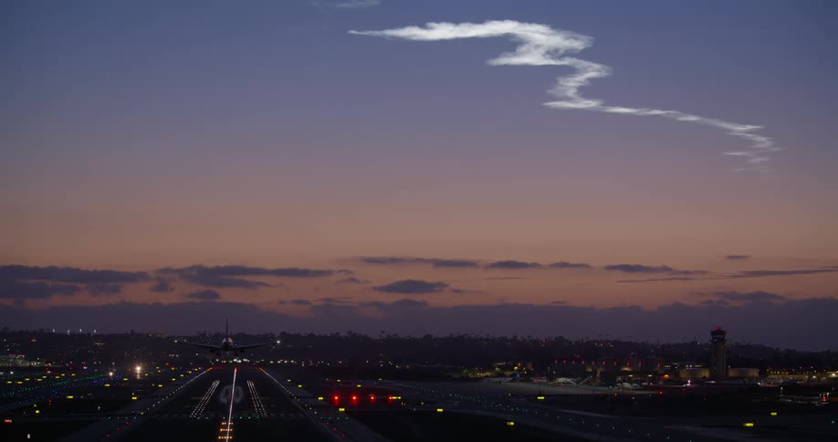 An airplane lands on an illuminated airport runway at dusk as a unique cloud formation hangs in the evening sky