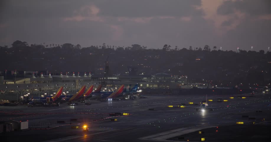 Airplanes taxi on a brightly lit runway at a bustling airport during a cloudy twilight