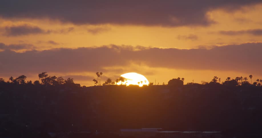 A beautiful golden sun sets behind a silhouetted hill with palm trees under a cloudy sky