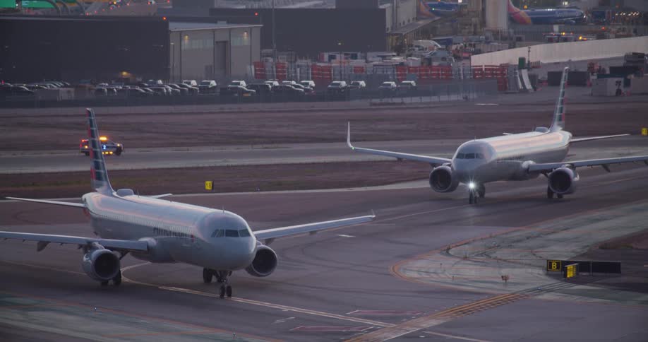 Two commercial airplanes wait on the tarmac at dusk with their lights flashing as a police car drives by in the background