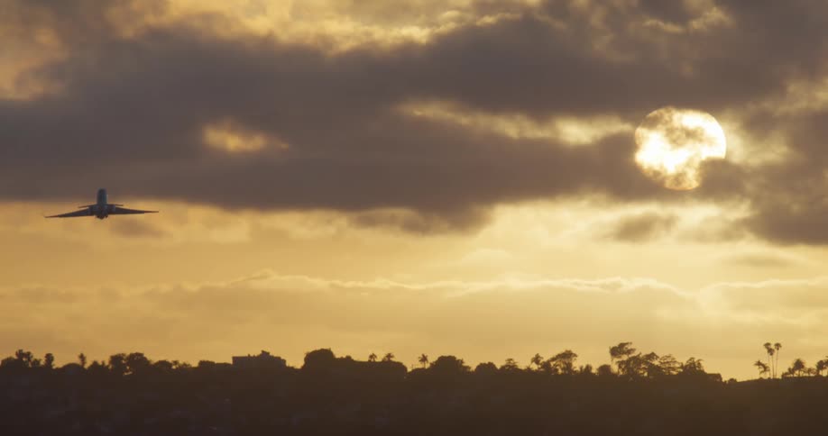A commercial airliner takes off and flies across the frame silhouetted against a dramatic golden sunset sky