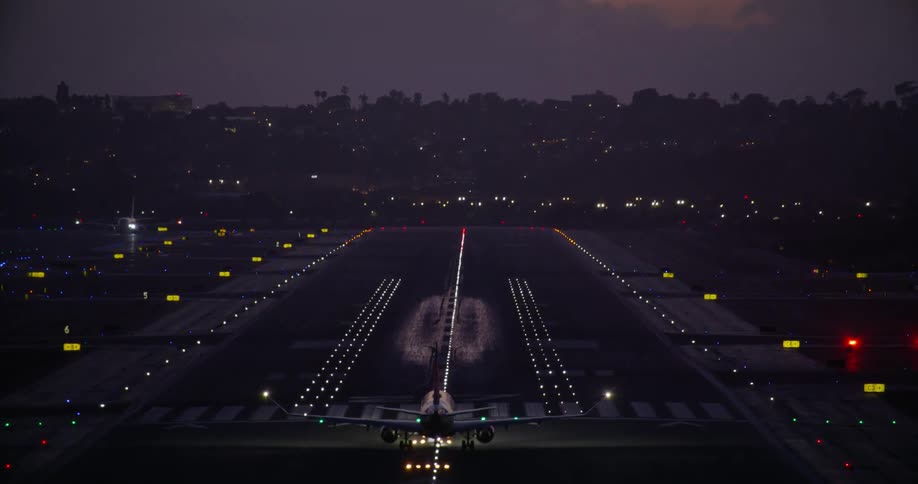 A passenger jet accelerates down an illuminated airport runway at night and takes off into the twilight sky