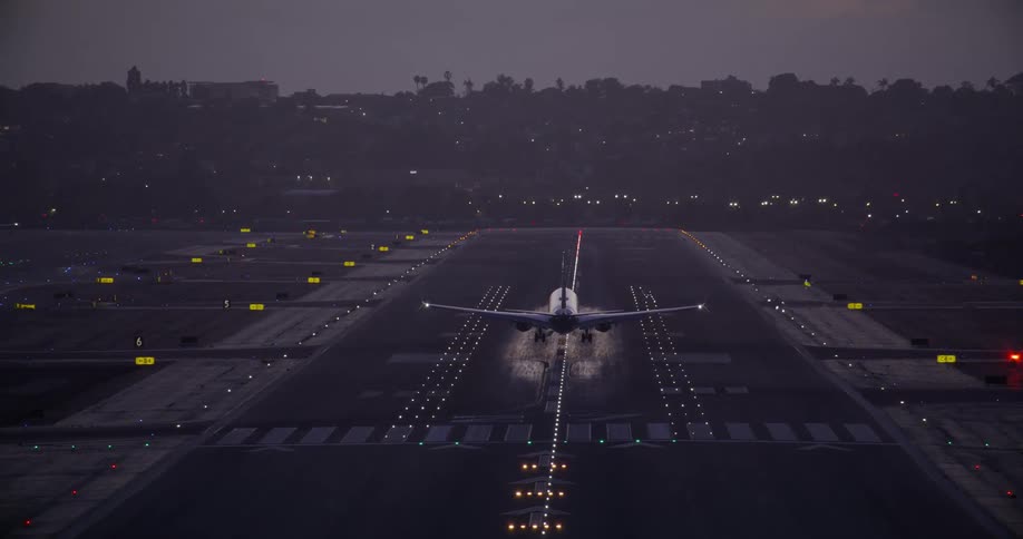 A passenger jet is seen from behind as it descends and lands on a brightly lit runway at dusk