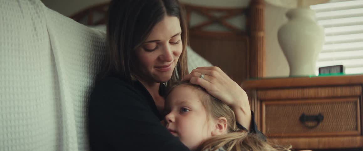 A loving mother gently holds and comforts her young daughter in a quiet, sunlit room