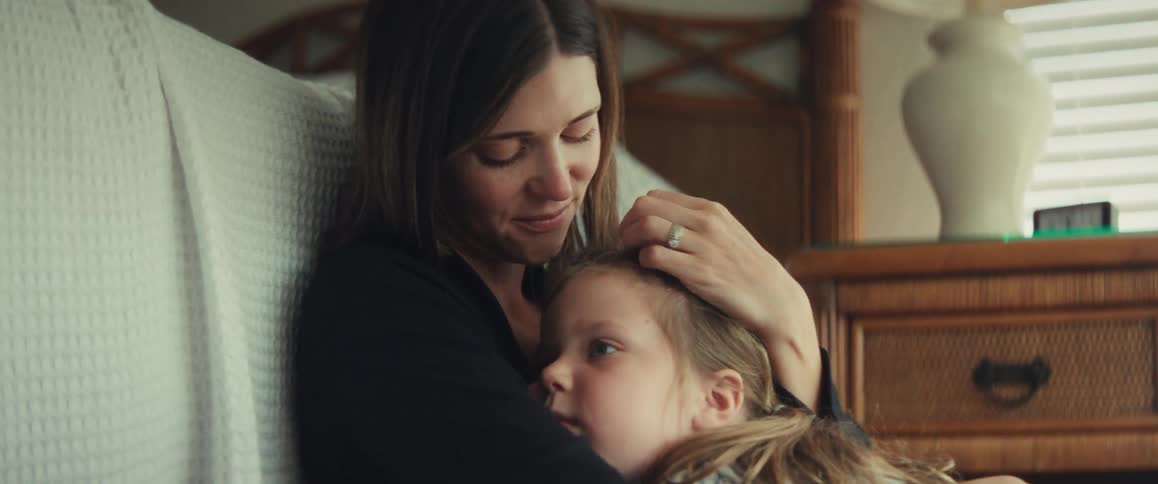 A loving mother gently holds and comforts her young daughter in a quiet, sunlit room